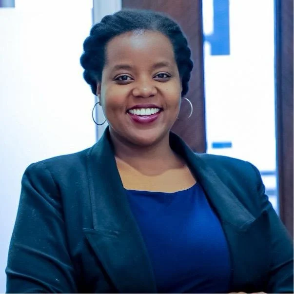 A woman with dark hair styled in braids, wearing hoop earrings, a dark blue top, and a black blazer, smiling.