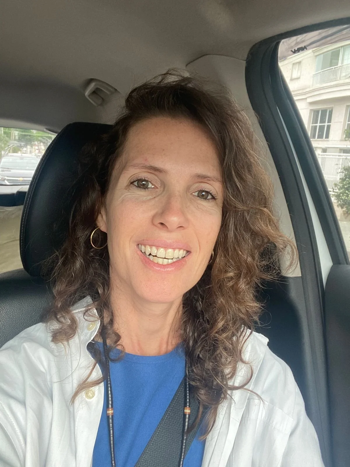 A woman with curly brown hair and hoop earrings smiling while sitting in a car.