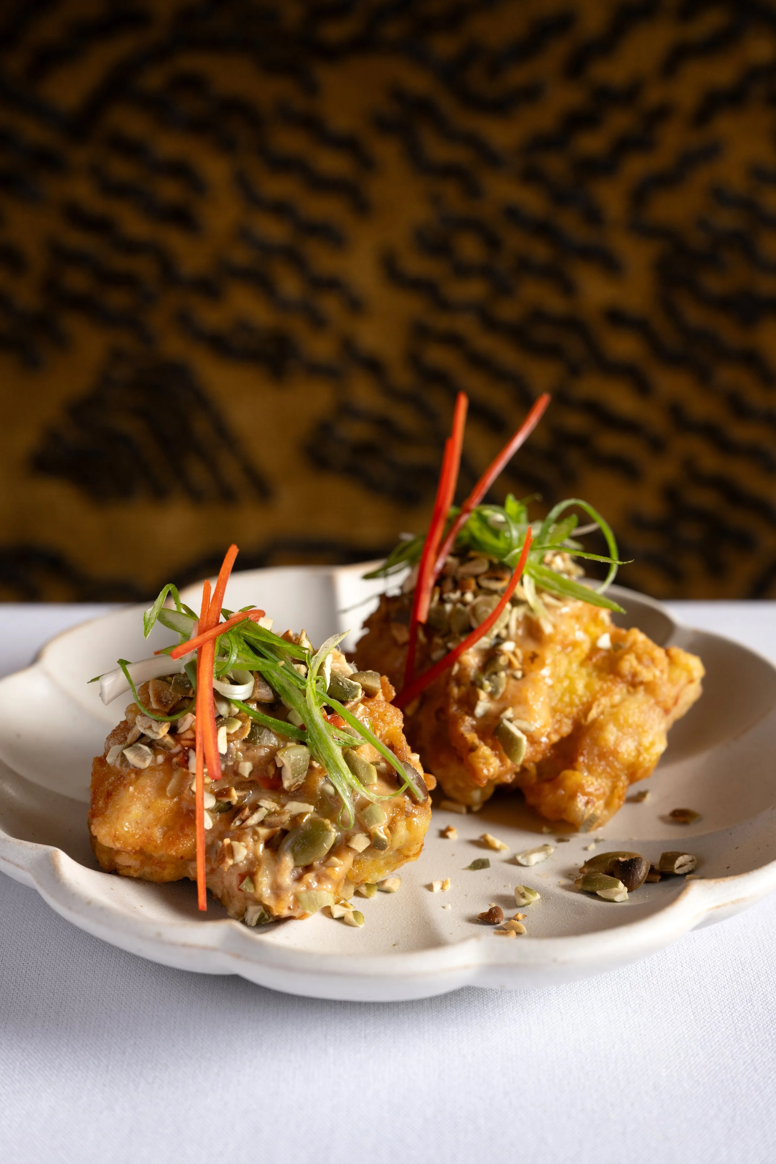 Close-up of two fried pieces of food topped with chopped green herbs, shredded carrots, sunflower seeds, and sliced almonds on a white plate.