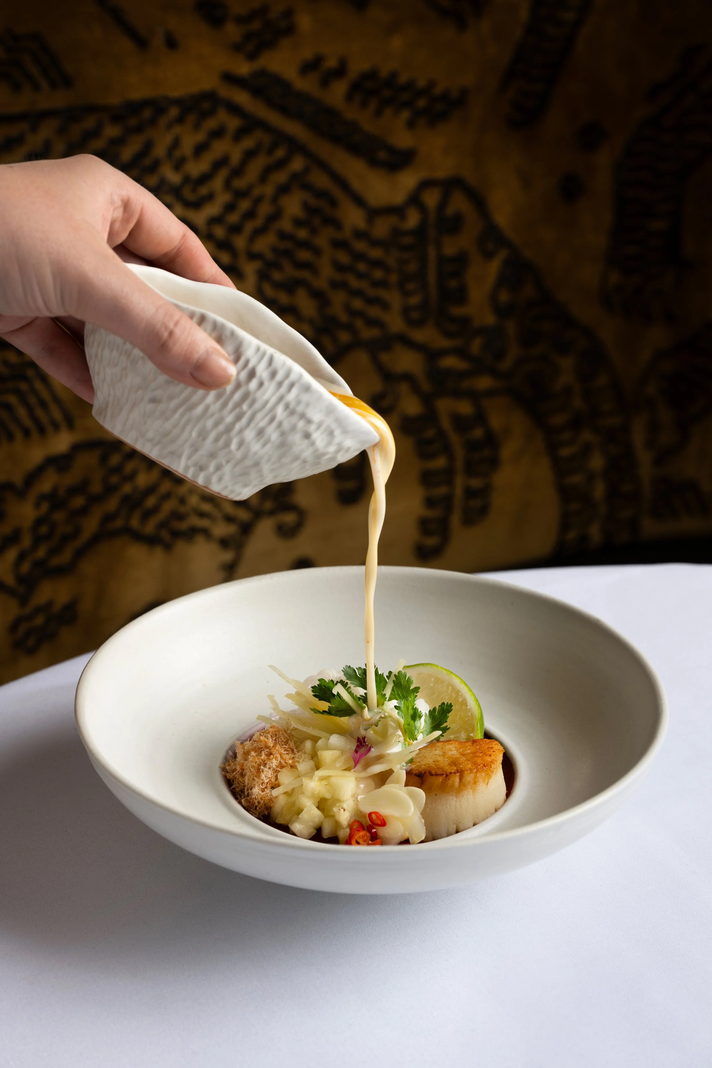 A hand pouring creamy sauce over a bowl of gourmet seafood dish with scallops, garnished with lime, cilantro, and red pepper slices against a patterned background.