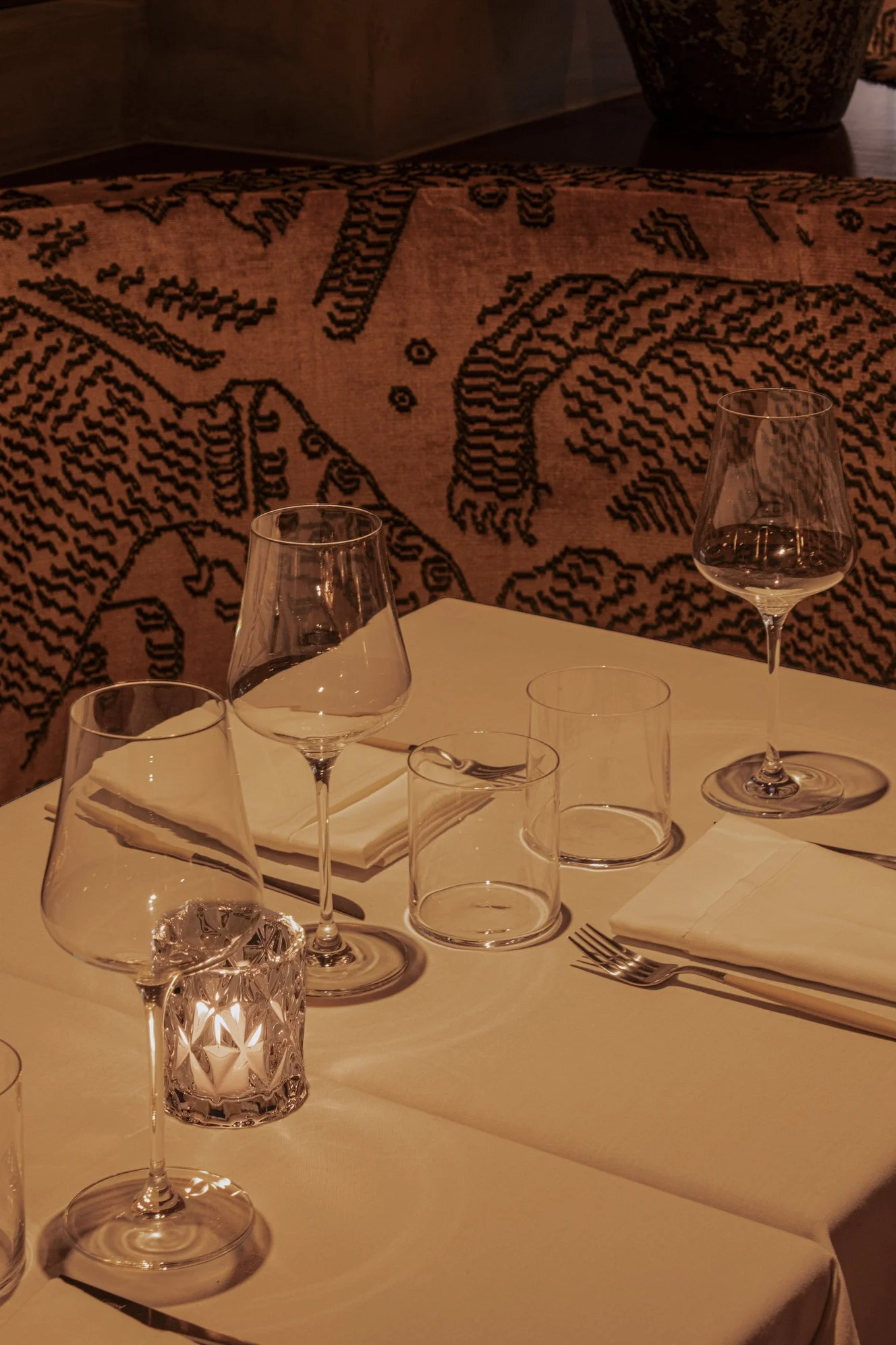 A table set with multiple empty wine glasses, water glasses, white napkins, and silverware, with a patterned fabric backdrop and a lit candle in a decorative holder.