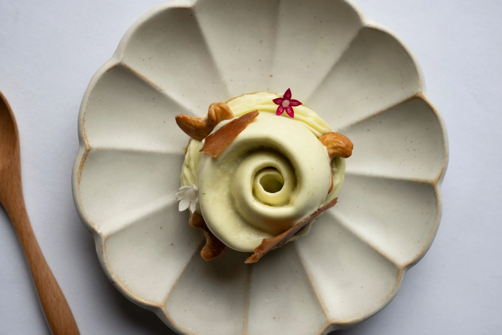 A gourmet dessert on a ceramic plate with petal-shaped sections, decorated with a pink flower, nuts, a white chocolate garnish, and a small flower, with a wooden spoon beside it.