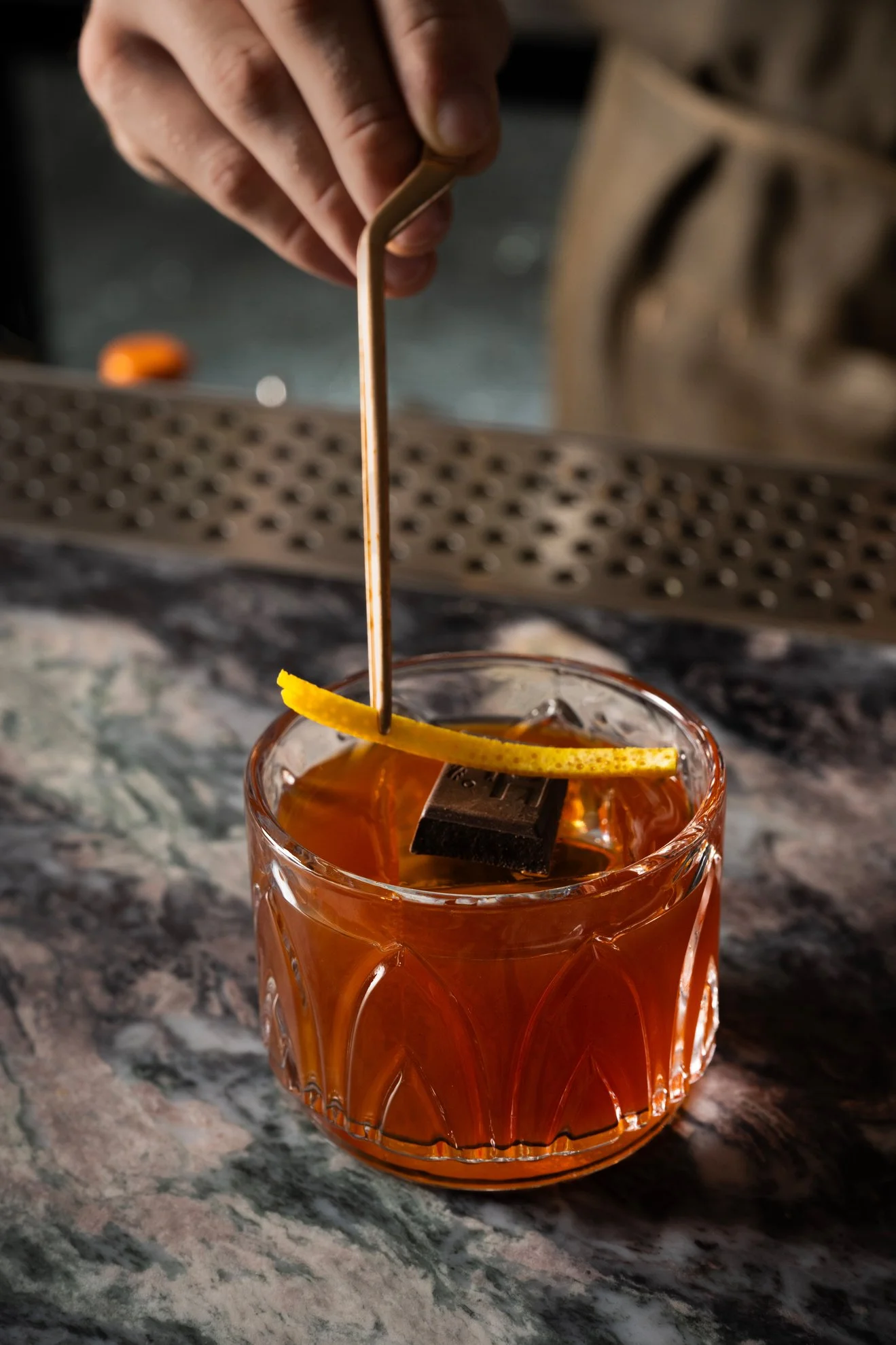 Close-up of a hand holding a cocktail pick with an orange peel twist, inside a glass with an amber-colored beverage on a marble countertop.