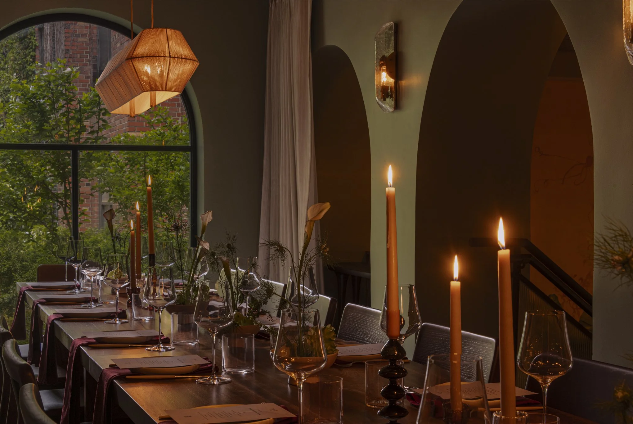 Interior of a restaurant with a long table set for dining. The table has multiple wine glasses, napkins, and small flower arrangements. Candles are lit in glass holders, and a large arched window reveals greenery and a brick building outside. A penda