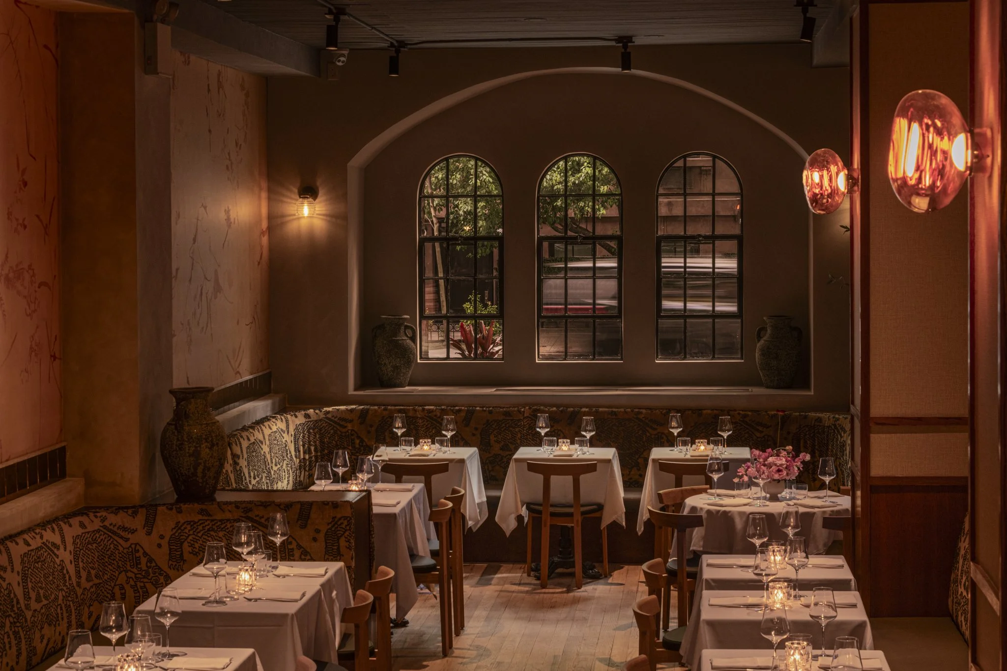 Interior of a restaurant with white tablecloths, wine glasses, pink flowers, and warm lighting, including large arched windows showing outdoor greenery.