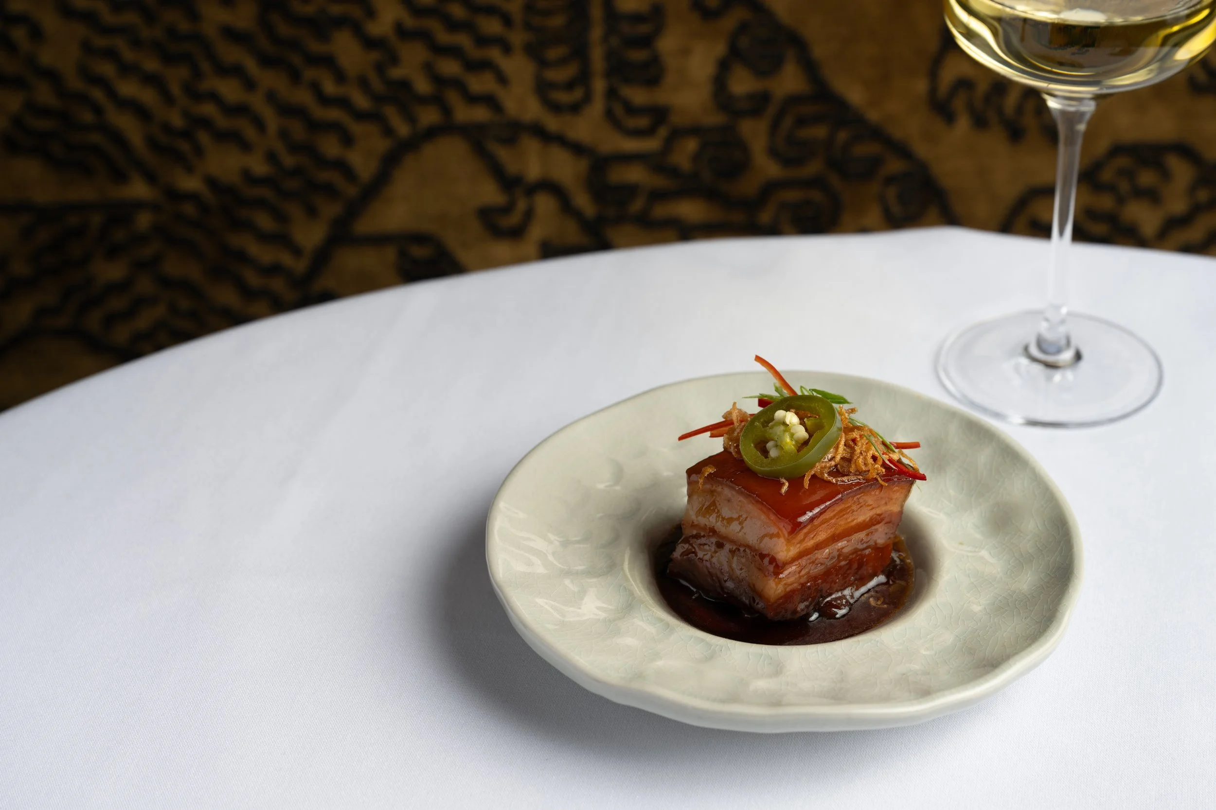 A small bowl of sliced, glazed fish topped with green chili slices, shredded red pepper, and crispy garnish, placed on a white tablecloth with a glass of white wine nearby and a patterned background.