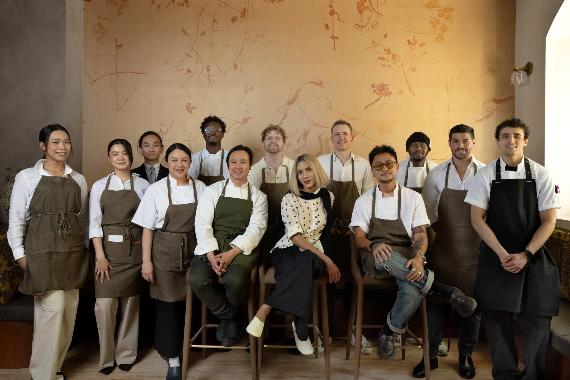 A diverse group of fifteen people, including women and men of various ethnicities, are posing for a photo in a restaurant or cafe. Some are standing while others are seated. They are all dressed in professional or casual attire, with some wearing apr