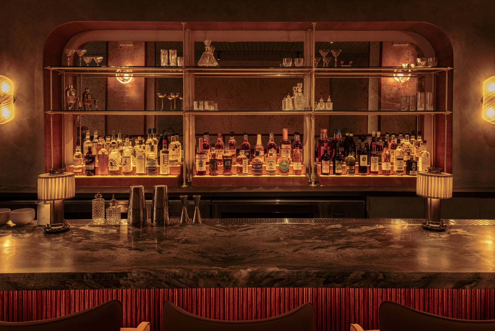 A bar with an array of various liquor bottles illuminated by warm lighting behind a marble countertop.