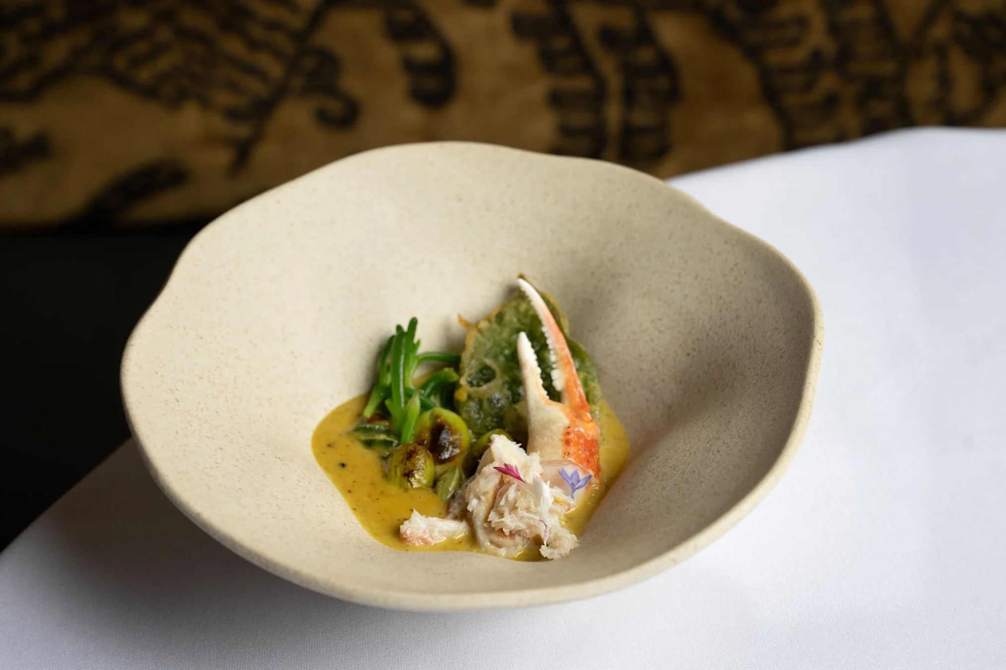 A white bowl with a seafood dish including crab, green vegetables, and a yellow sauce garnished with edible flowers.