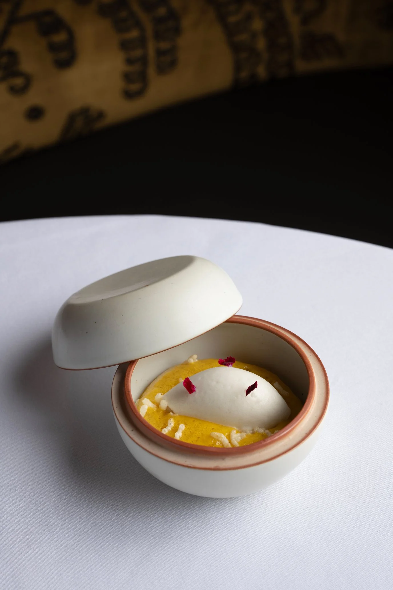 A small, open ceramic bowl containing a dessert with a dollop of white cream, yellow custard, and red sprinkles, set on a white surface with a partially visible egg-shaped object and a lid nearby.