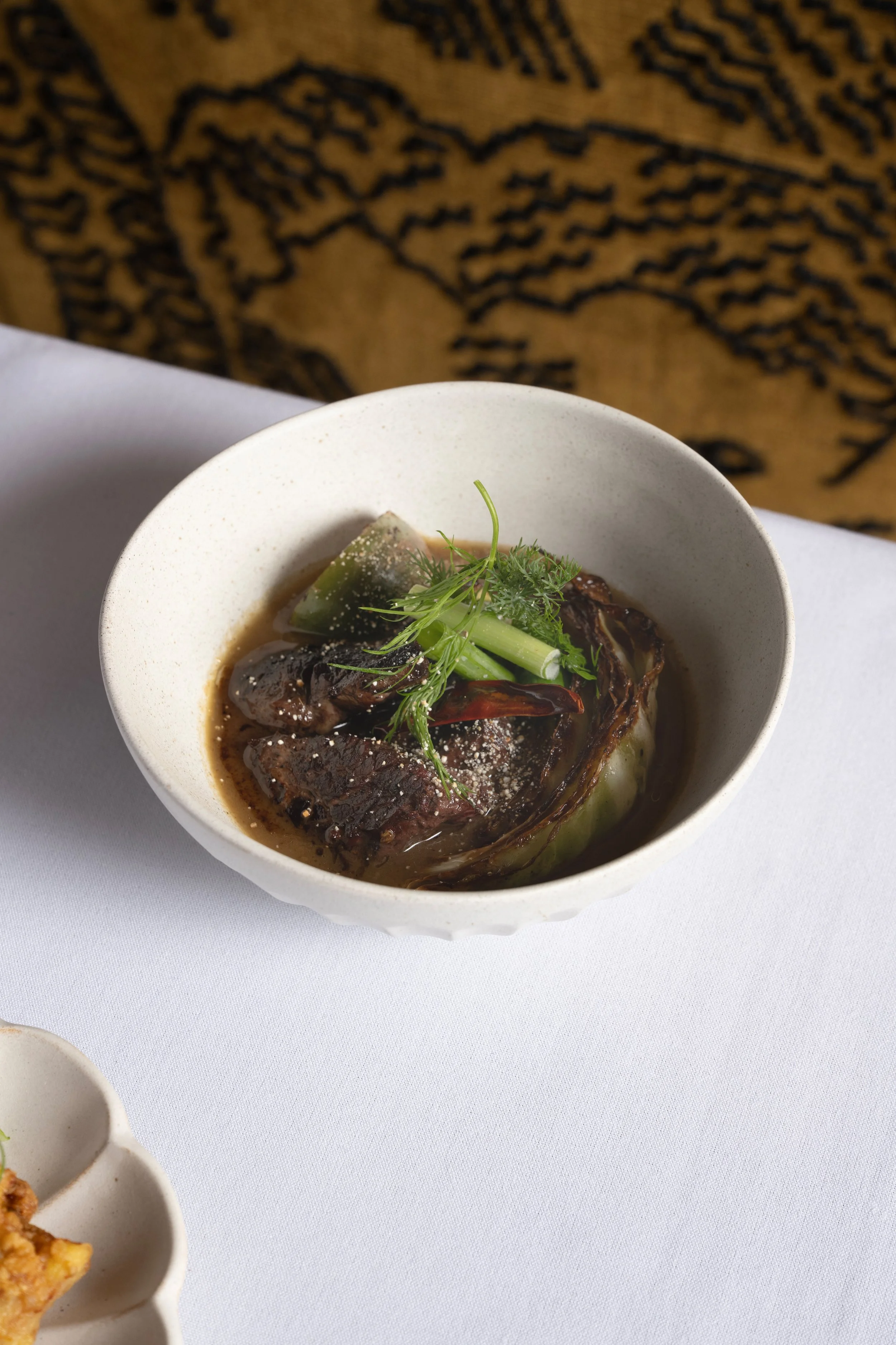 A bowl of beef stew garnished with green herbs on a white tablecloth, with part of a white dish in the lower left corner and a printed fabric in the background.