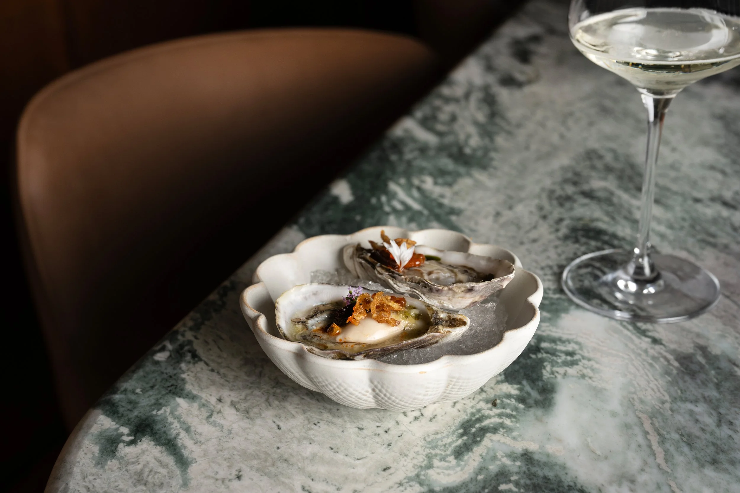 A white scalloped dish with raw oysters topped with garnishes sits on a green marble countertop. In the background, a glass of white wine is partially visible.