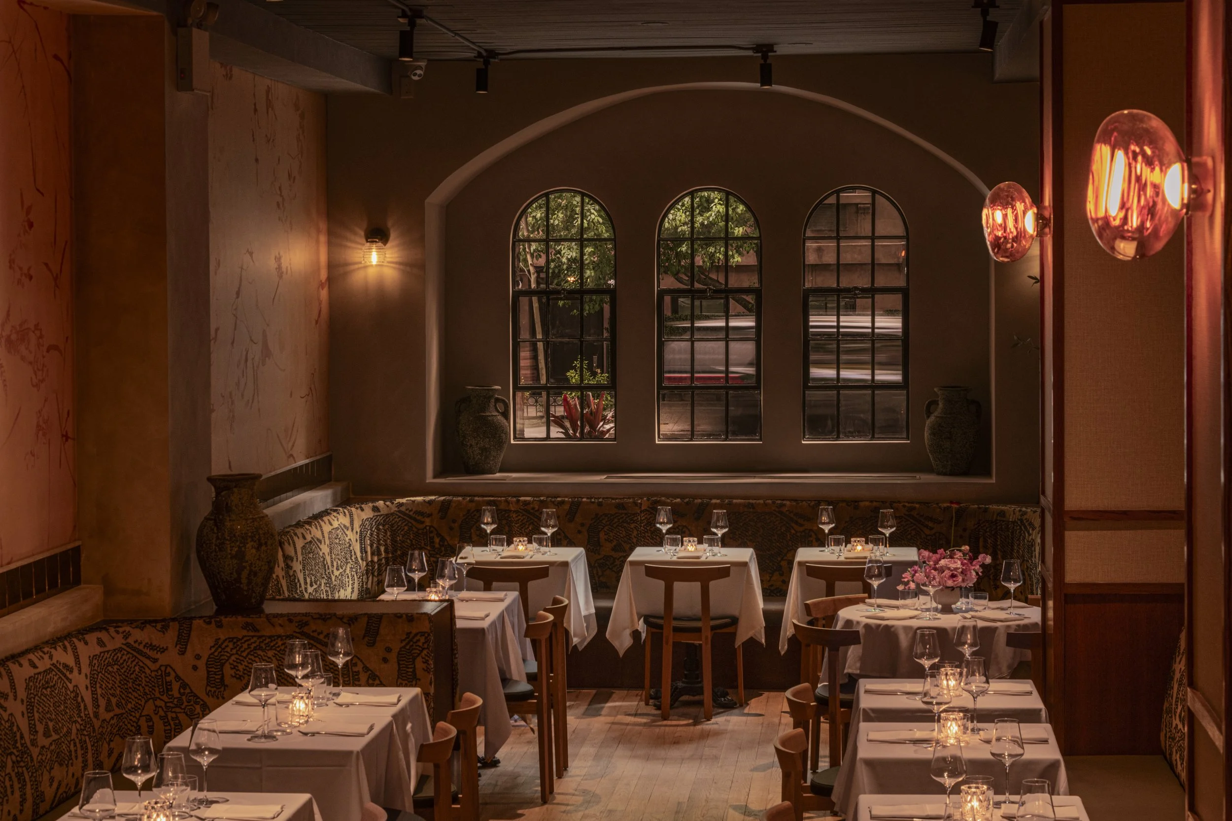 A cozy restaurant interior with large arched windows, wood accents, and warm lighting. Dining tables with white tablecloths, wine glasses, and floral centerpieces are arranged throughout the space.