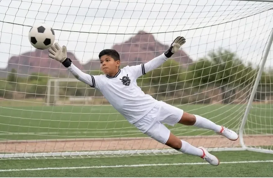 A young soccer goalkeeper in a white uniform with black accents making a diving save in front of a goal, with a soccer ball near his outstretched hands on a grassy field.