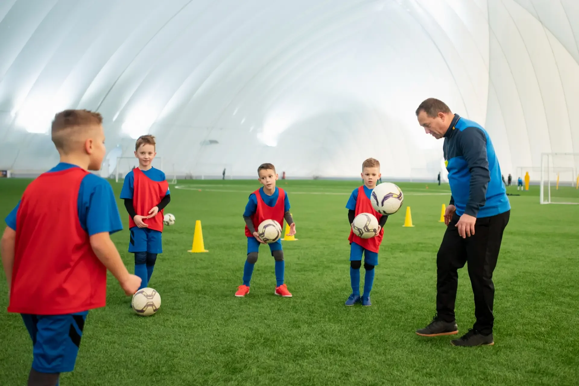 A coach instructs young children during soccer practice inside a large indoor facility. The children are holding soccer balls and are dressed in blue and red training vests and shorts. Cones are set up on the field for drills.