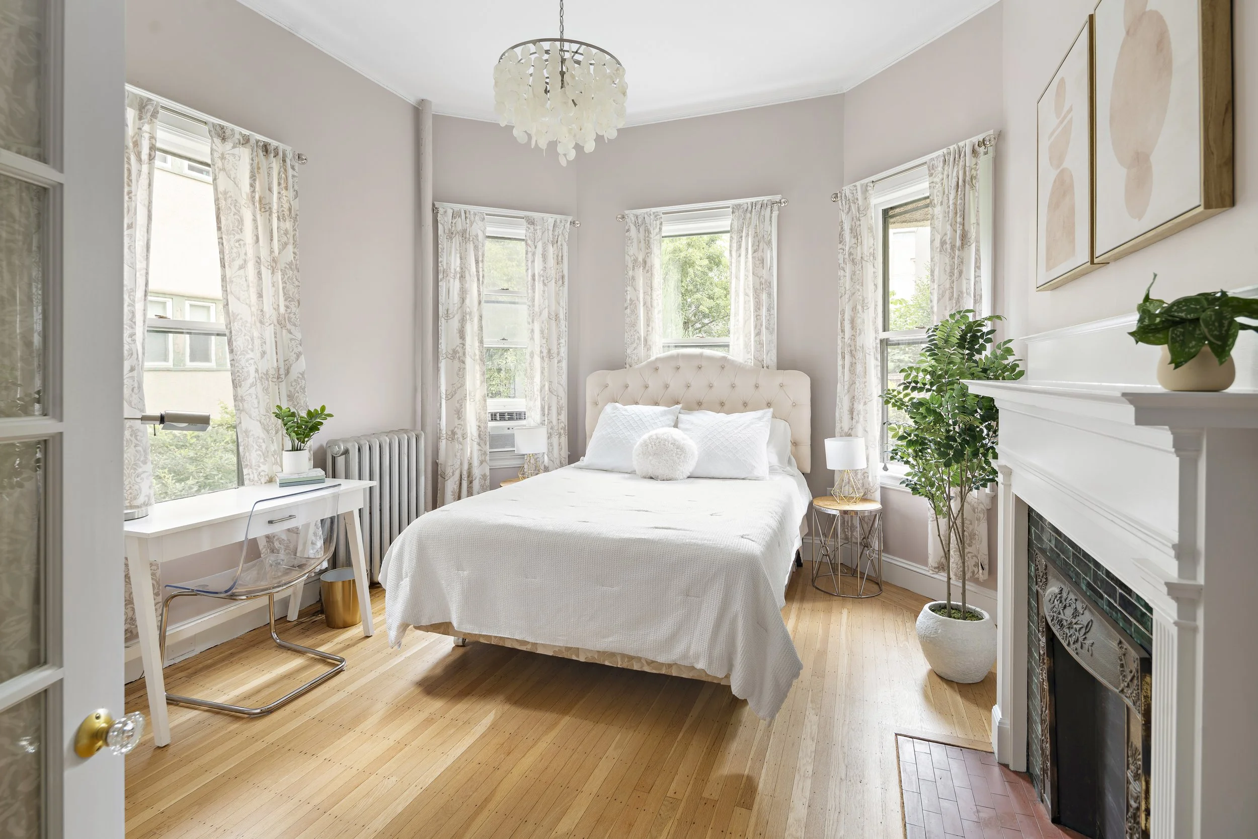 Bright bedroom with a cream upholstered bed, white bedding, and two bedside tables with lamps. Floral curtains frame three windows, with a white fireplace on the right and a small desk with a clear chair on the left, decorated with green potted plant