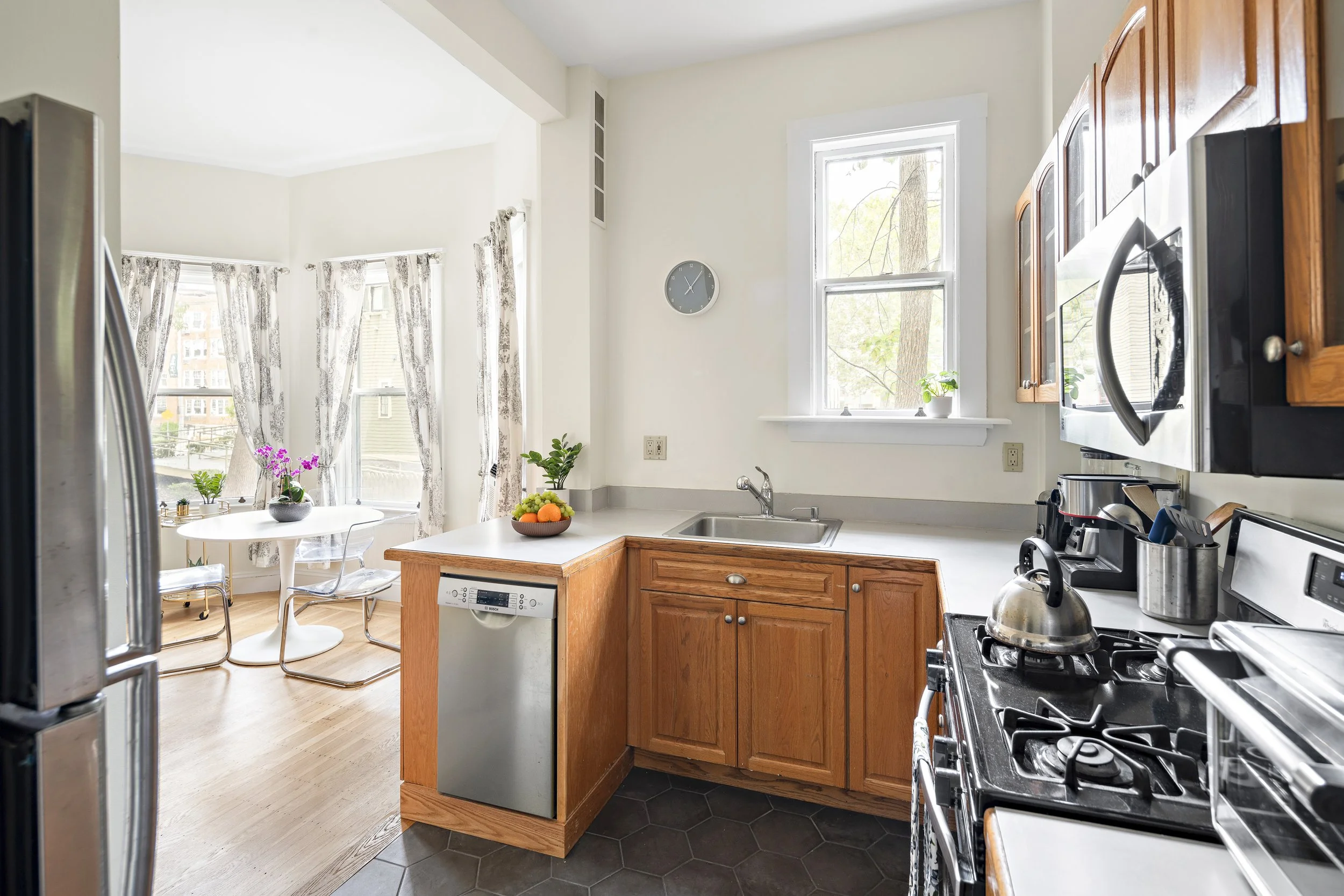 Kitchen with wooden cabinets, stainless steel appliances, a window with a view of trees, a small fruit bowl on the counter, and a dining area with a round table and chairs near a bay window with floral curtains.