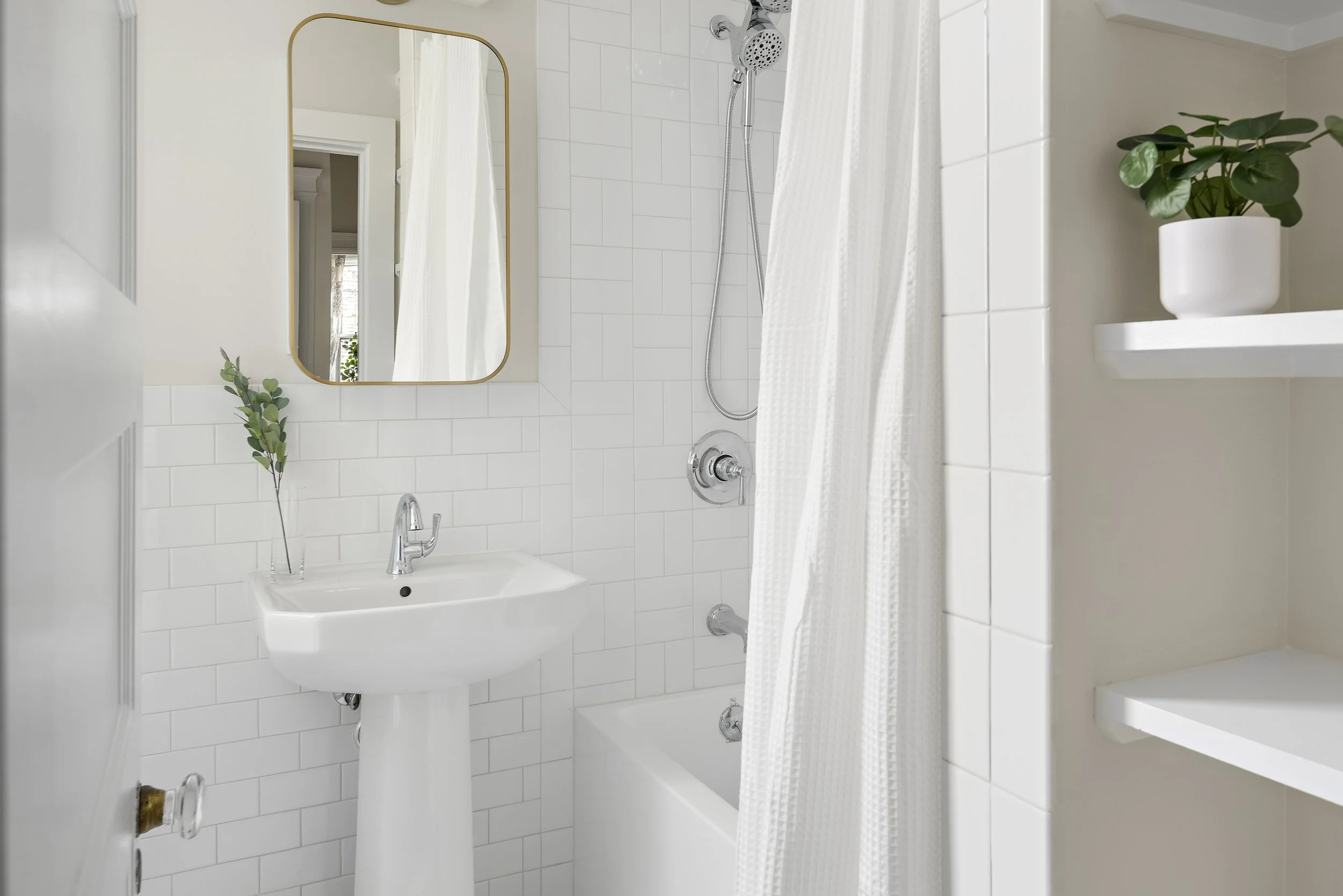 A clean, minimalistic bathroom with a white pedestal sink, a small mirror, a shower with a curtain, and a white shelf with a potted plant.