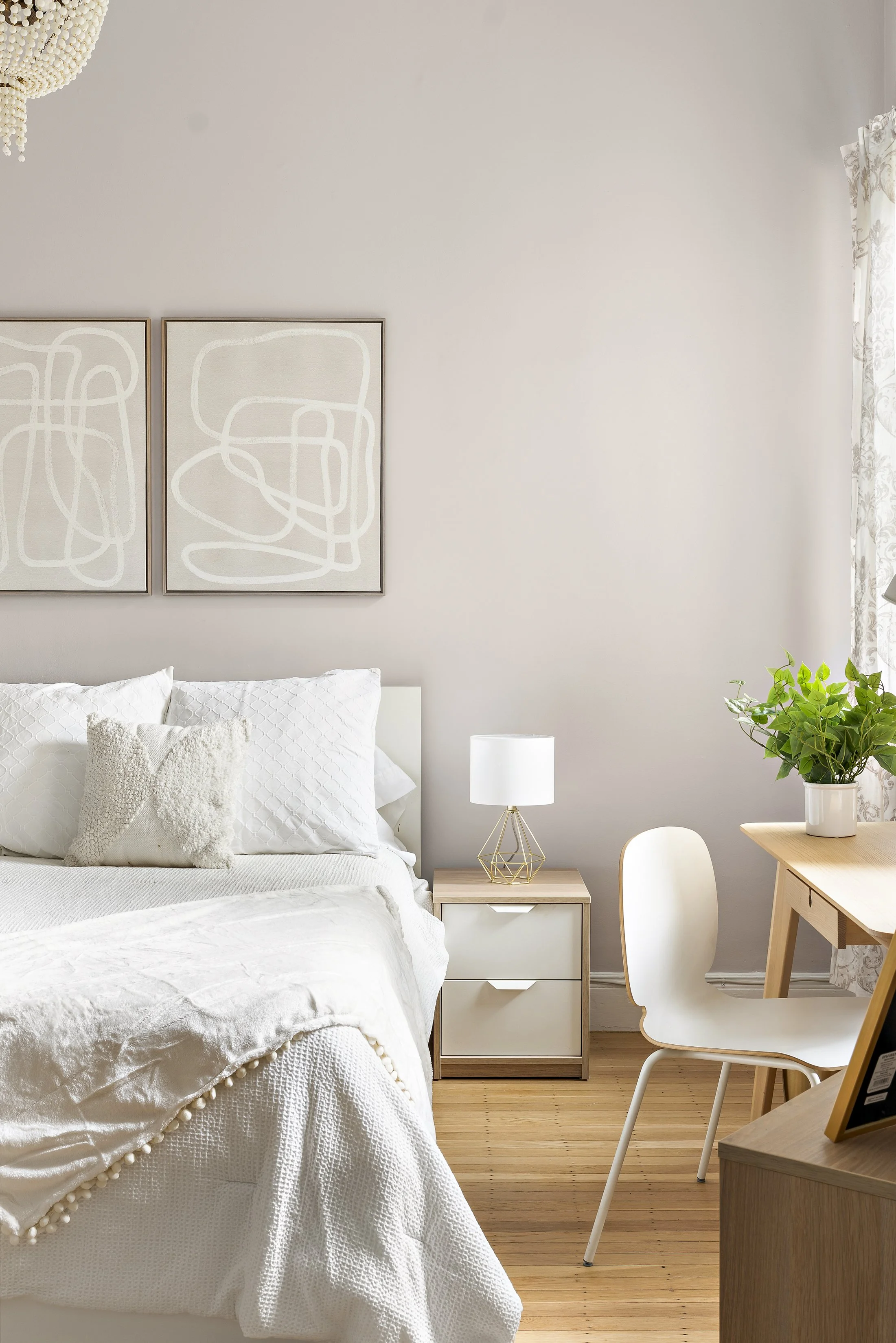A bright, minimalist bedroom with a white bed, abstract art on the wall, a bedside table with a modern lamp, a wooden desk with a white chair, and a green plant in a white pot.
