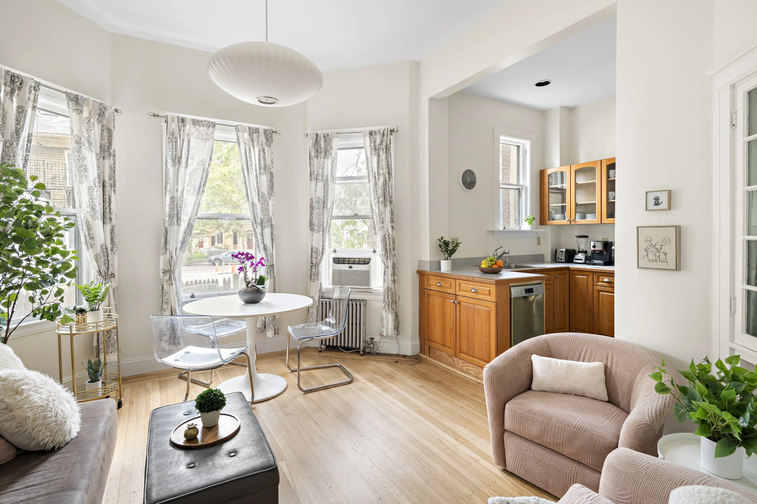 Bright living room with white walls, wooden flooring, a pink armchair, a beige sofa with cushion, a small round dining table with transparent chairs, and a kitchen with wooden cabinets and small window, decorated with plants and flowers.