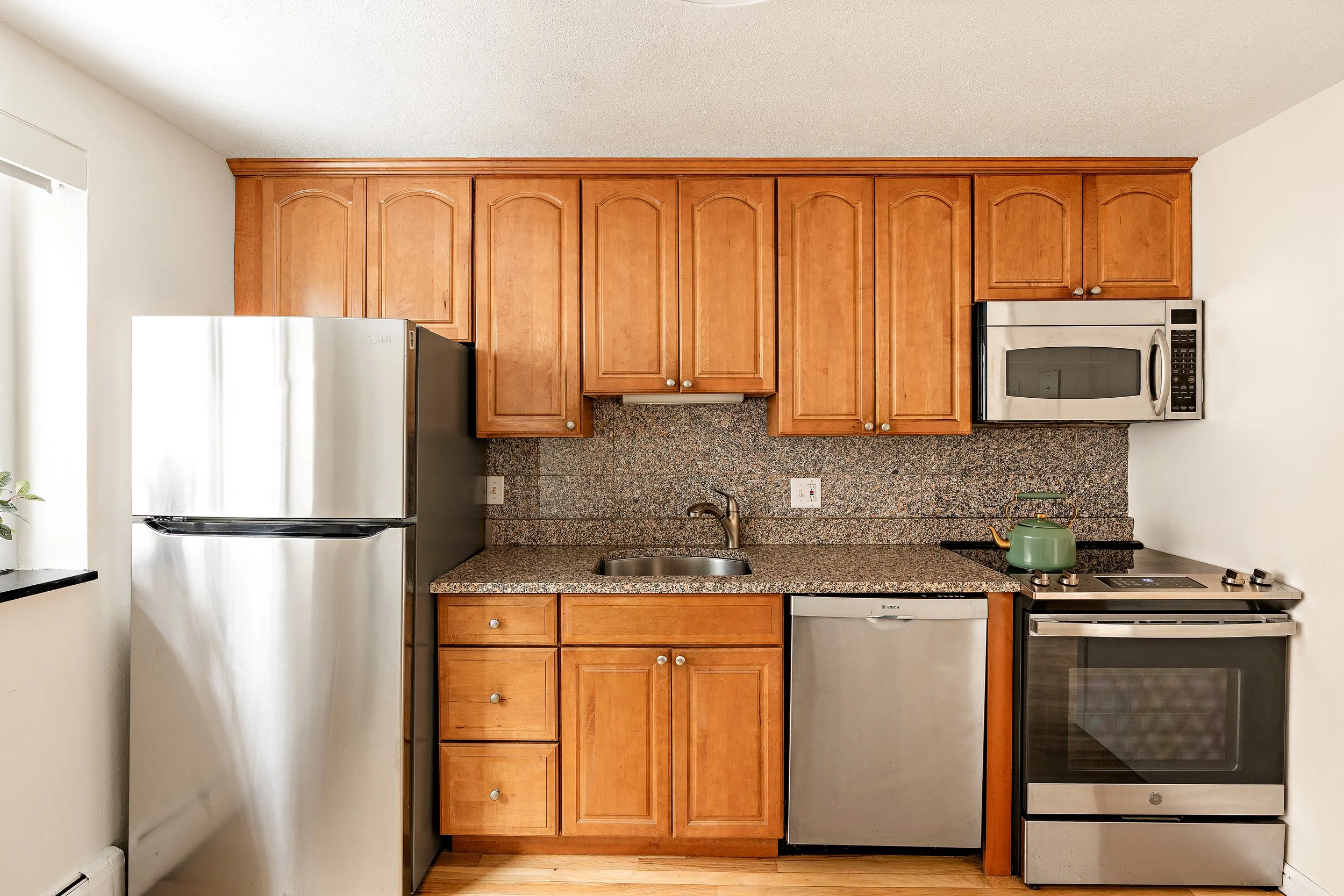 A compact kitchen with light wood cabinets, a granite countertop, stainless steel refrigerator, microwave, dishwasher, stove with a green kettle, and a small window on the left side.