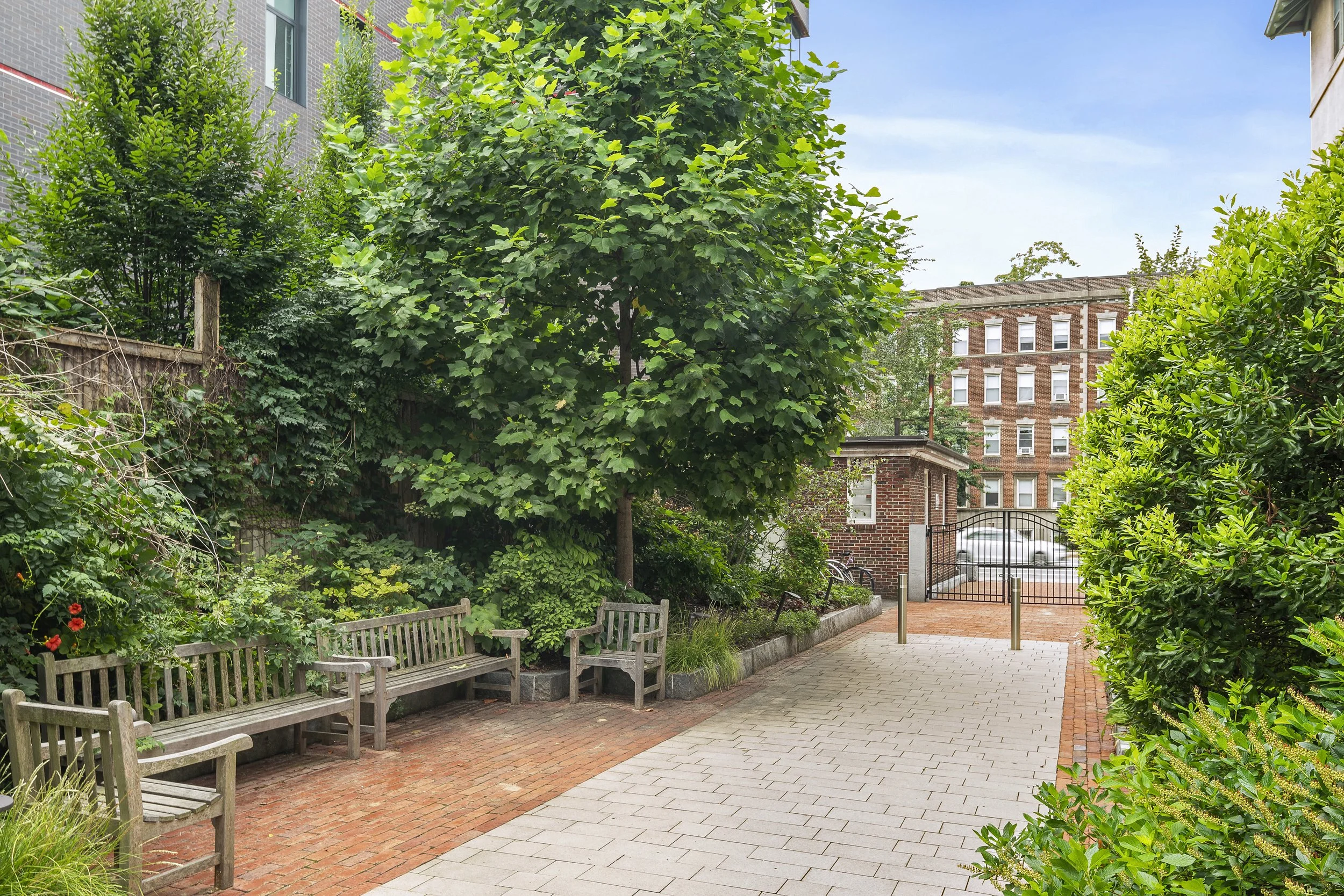 A tranquil urban courtyard with wooden benches, lush green trees, and brick-paved pathways surrounded by commercial buildings, secured by a gate.
