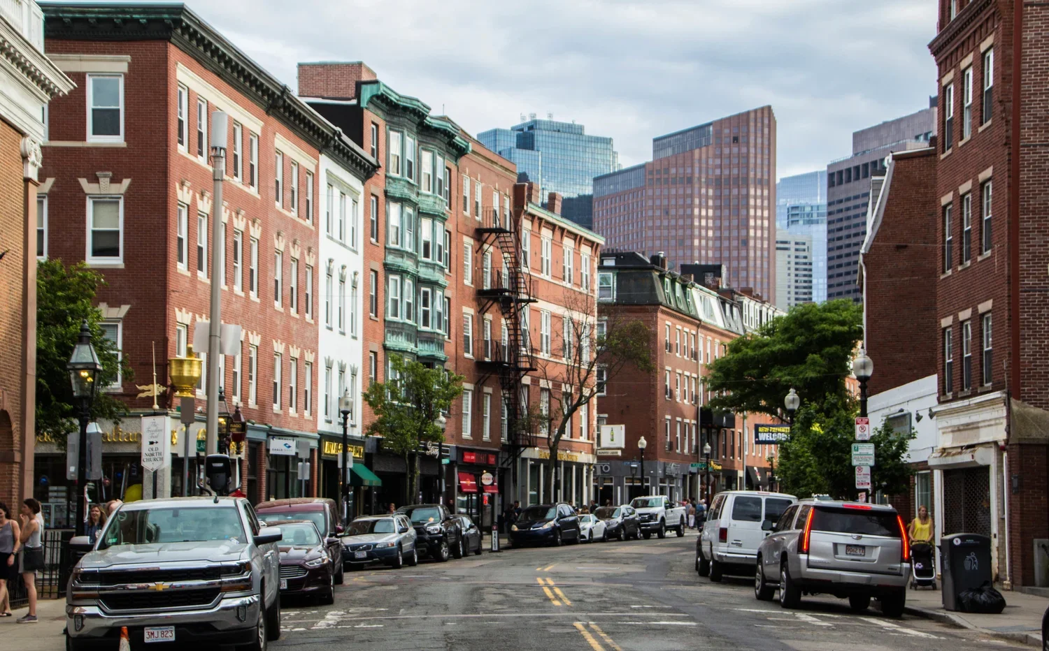 City street with parked cars, pedestrians, brick buildings, trees, and modern skyscrapers in the background.