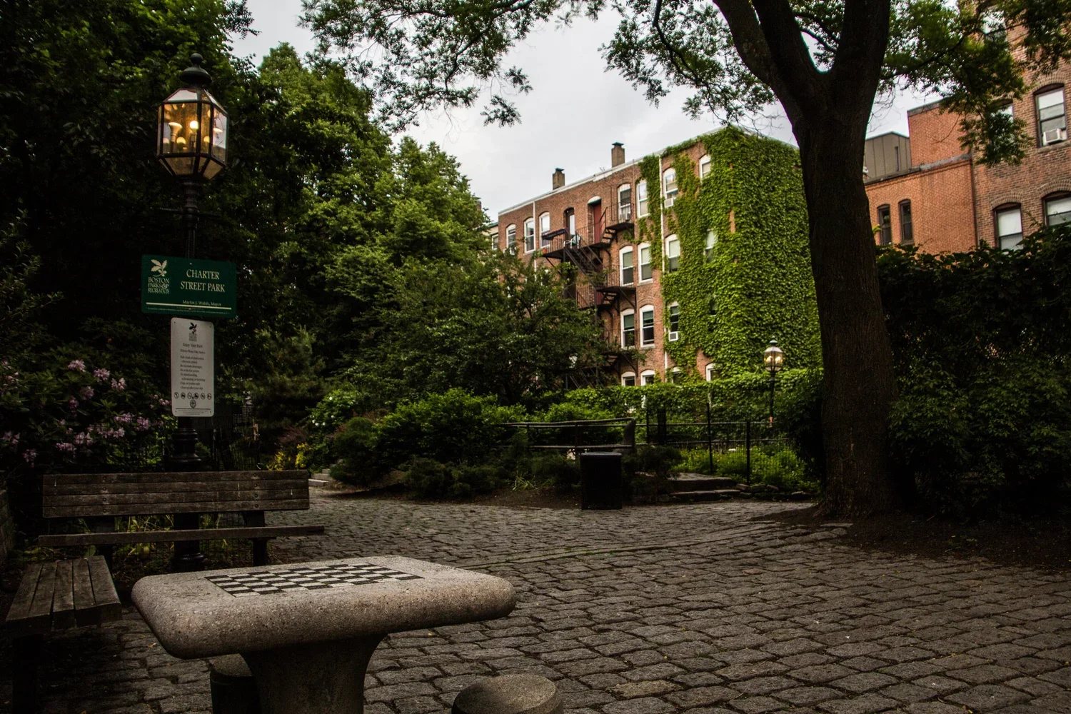 A small park with a cobblestone pathway, benches, lampposts, trees, and a brick residential building with ivy on the exterior surrounding the park.