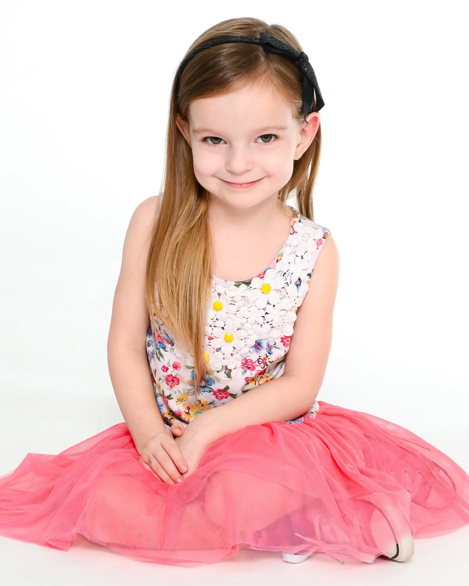 A young girl with long red hair, wearing a black headband, a floral sleeveless top, and a pink tutu, sitting on the floor against a plain white background.