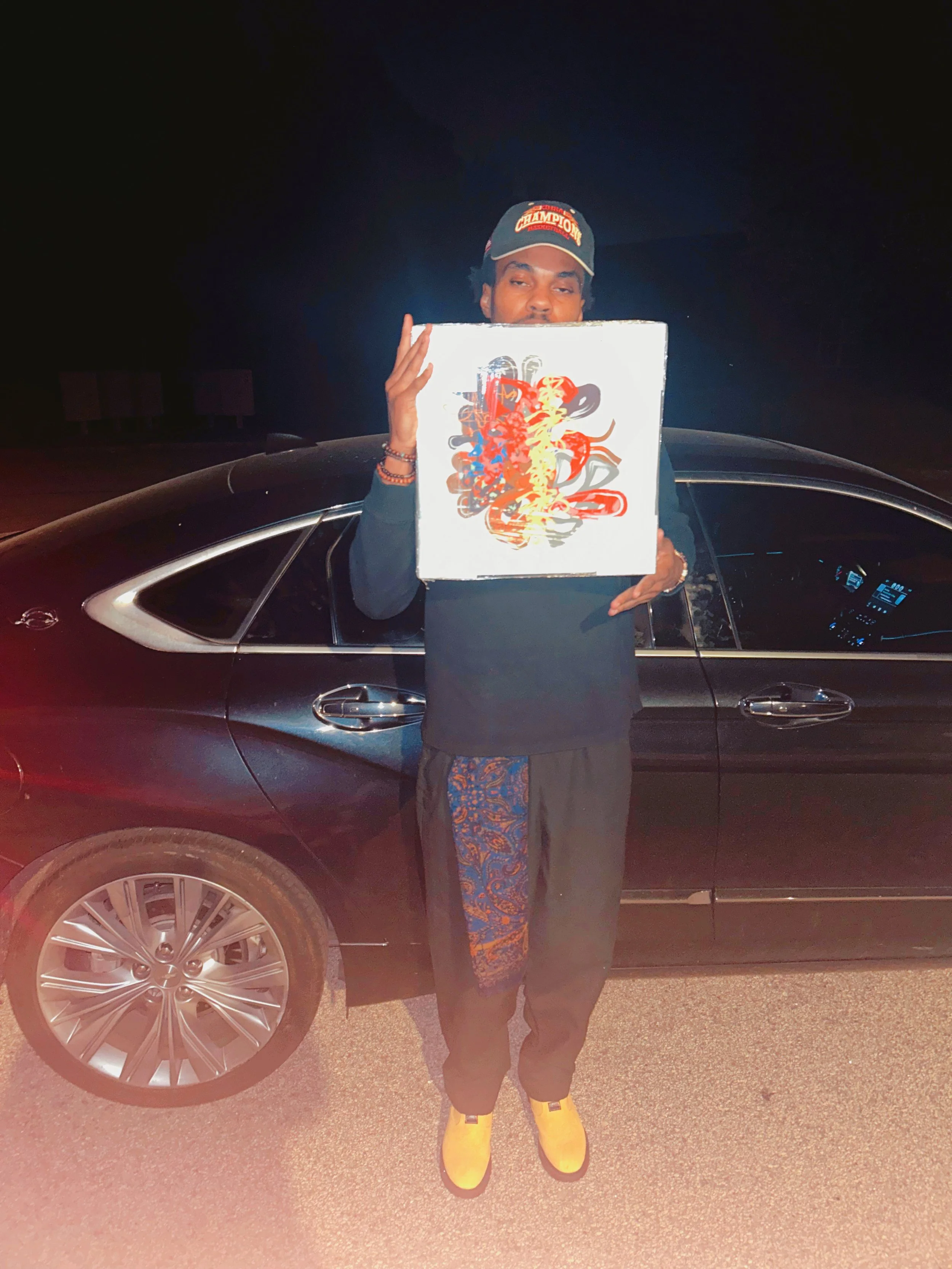 Person standing in front of a black car, holding a colorful abstract art piece, wearing a baseball cap, black pants, and bright yellow shoes at night.
