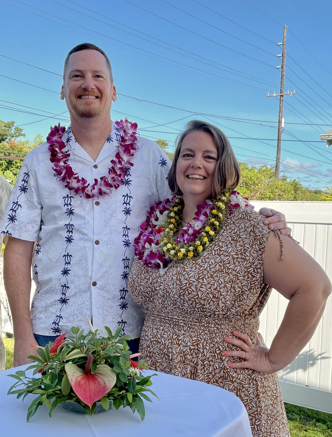 Jennifer Tobey and husband smiling outdoors, wearing Hawaiian shirts and leis, standing near a table with a floral arrangement, with power lines and trees in the background.