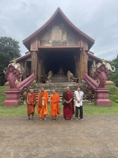 Five men, three in orange robes, one in maroon robes, and one in white casual clothing, stand in front of a traditional wooden temple with dragon statues on either side of the stairs.