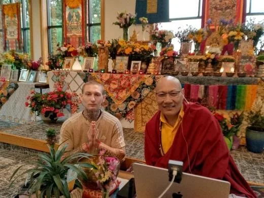 A young man with tattoos, holding his hands in a prayer position, sitting next to a smiling monk dressed in traditional robes, in front of a decorated altar with flowers, pictures, and spiritual items.