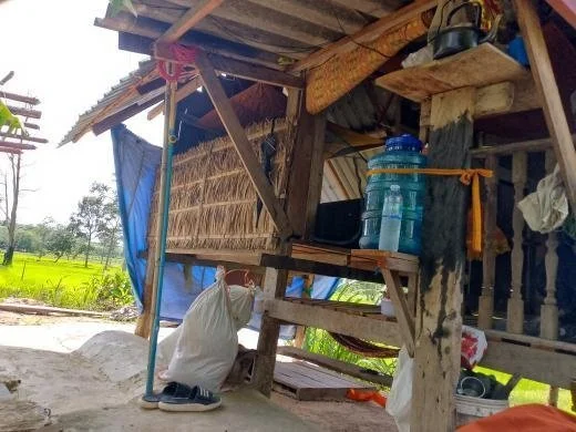 A person is crouching next to a makeshift wooden house with a water jug on a table, surrounded by lush green fields.