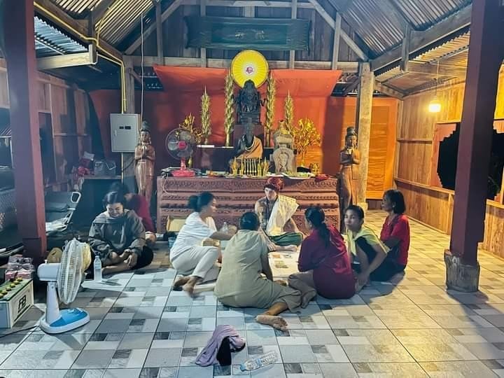 People sitting on the floor and participating in a religious or cultural ceremony inside a wooden building, with a shrine and statues in the background.