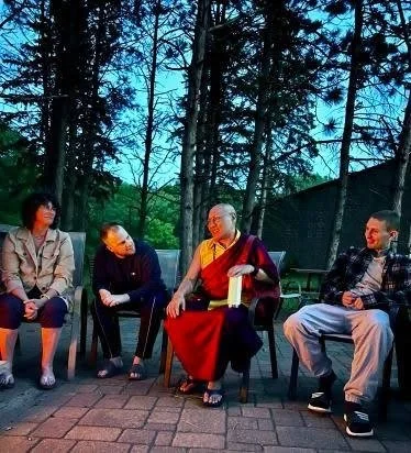 Four people sitting outside in a circle, trees in the background. One person is a Buddhist monk in traditional robes.