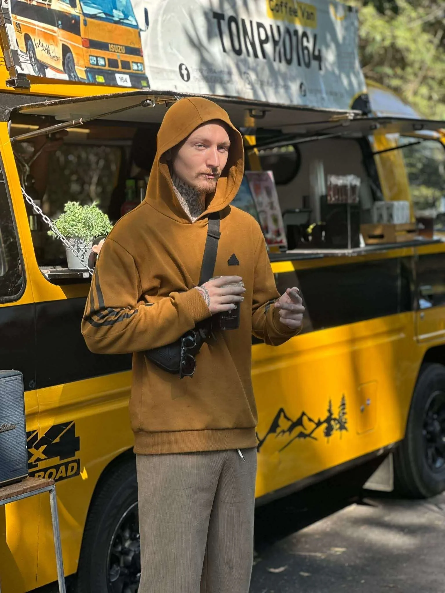 A man in a brown hoodie and pants standing in front of a yellow and black food truck, holding a drink.