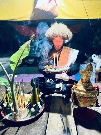 A man with a curly, light-colored wig blowing out candles on a birthday cake at a celebration, with traditional Japanese decorations and plants in the background.