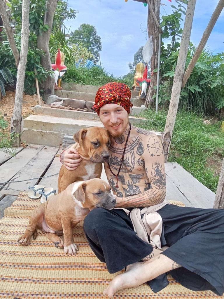 A man with tattoos, wearing a red patterned headscarf, sits on a woven mat outside with two puppies, one on his lap and one beside him. The background includes wooden steps, greenery, and decorative statues.
