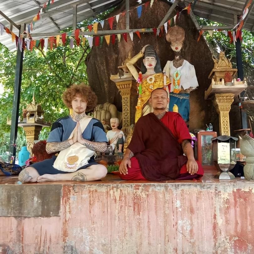 A Buddhist monk and a man with body tattoos sitting cross-legged on a platform in front of a large tree with Hindu or Buddhist statues and images, including a woman with a head covering and other small shrines, under a roof decorated with colorful fl