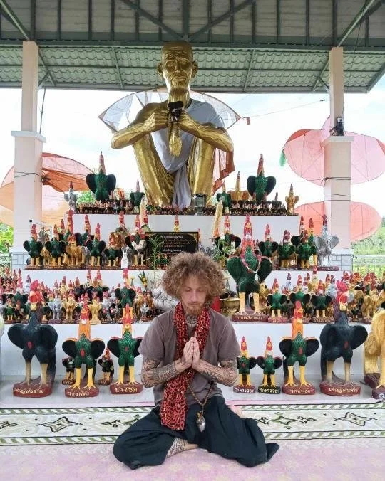 A person with curly hair, sitting cross-legged with hands in a prayer position, in front of a large golden statue of a sitting figure with multiple small animal statues surrounding it.