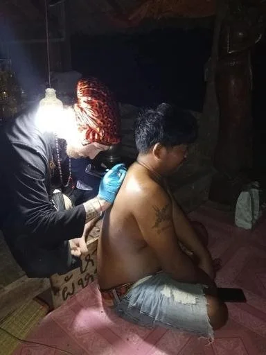 A man sitting on the floor getting a tattoo on his arm from a tattoo artist using a tattoo machine. The setting appears to be indoors with dim lighting.