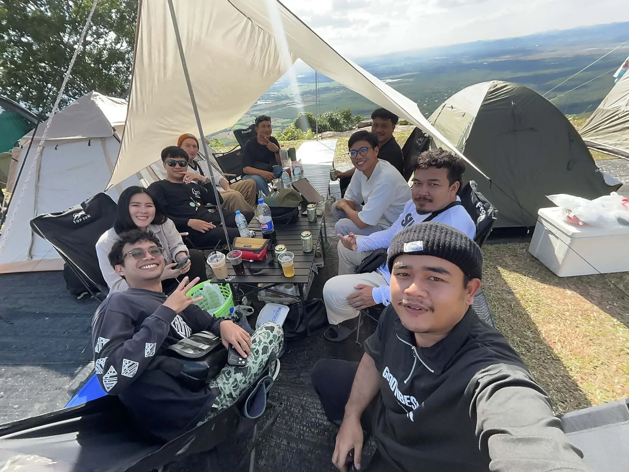 Group of friends sitting around a table outdoors under a canopy, enjoying drinks with tents and a scenic view of hills in the background.