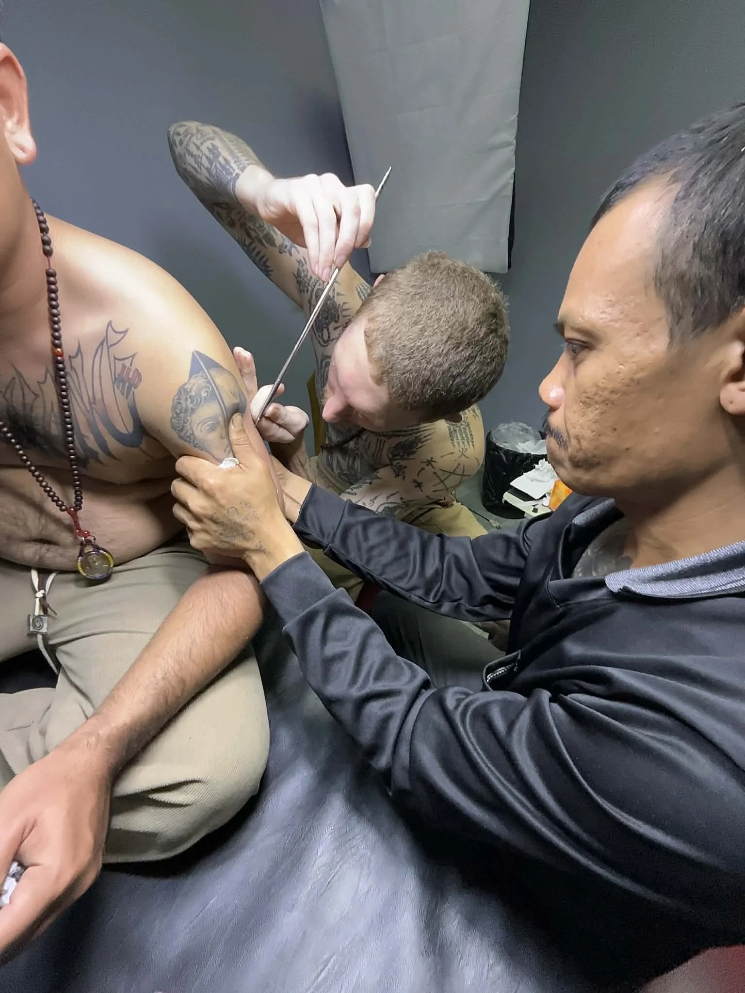 Man getting a tattoo on his chest from the tattoo artist, who is holding a tattoo needle, while another man assists.
