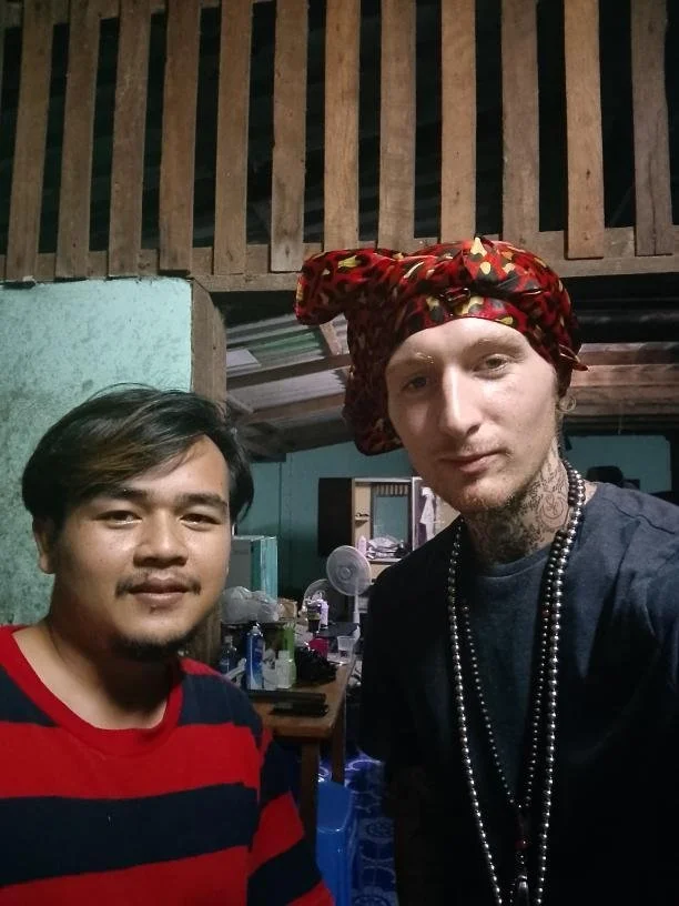 Two men taking a selfie indoors. The man on the left has black hair, a beard, and is wearing a red and black striped shirt. The man on the right has blond hair, tattoos on his neck, and is wearing a red headscarf with gold accents, a dark shirt, and 