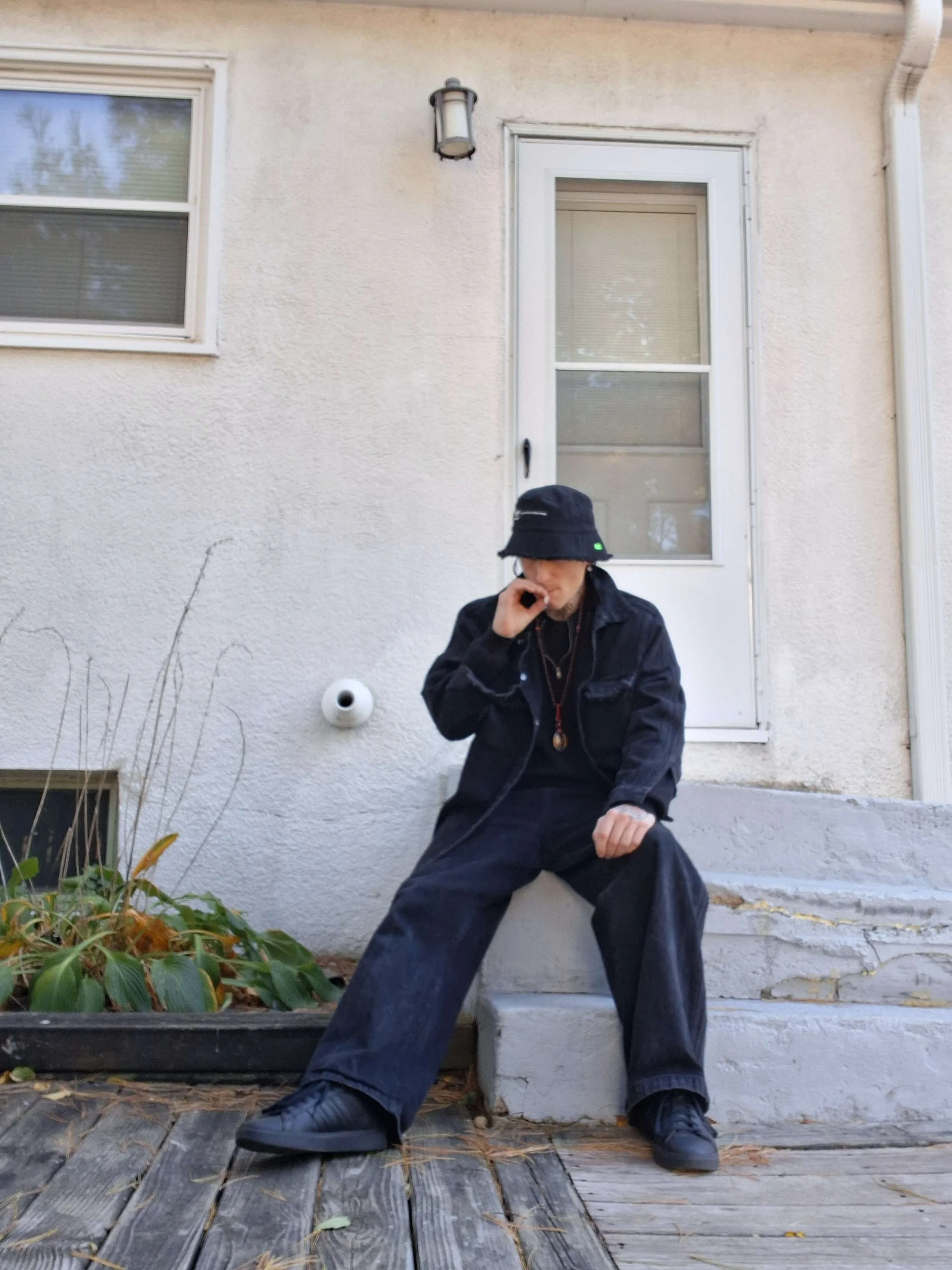 Person sitting on steps outside a house, wearing dark clothing, a bucket hat, and headphones, with a hand near their face, in front of a textured white wall and a window.