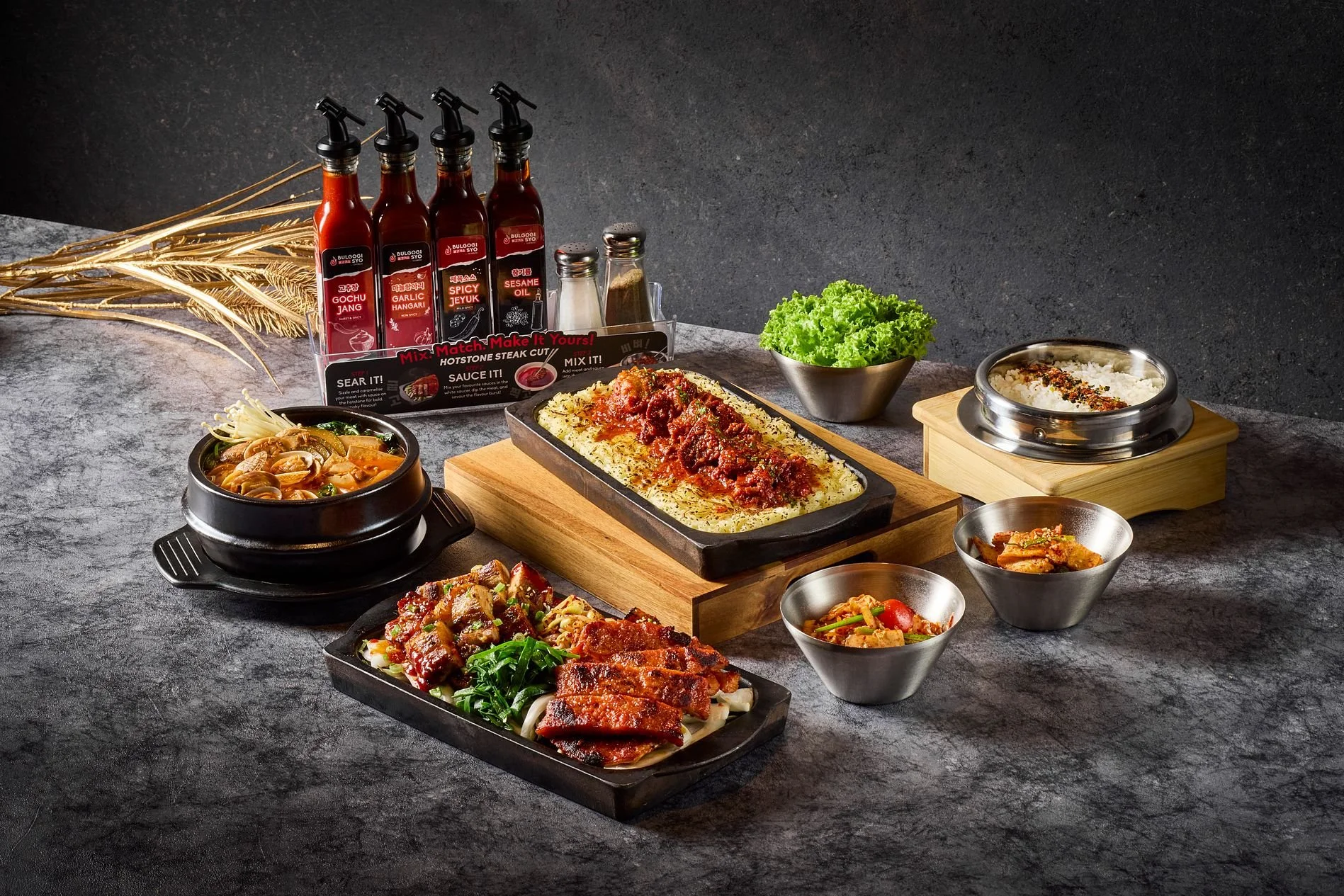 A variety of Korean dishes on a dark textured surface. Included are a tray with seasoned rice topped with red sauce, a bubbling hot stone pot with spicy soup, small bowls of vegetables and sauces, Korean condiments, a bowl of lettuce, and various side dishes. In the background, there are bottles of Korean sauces and condiments, and dried wheat stalks for decoration.