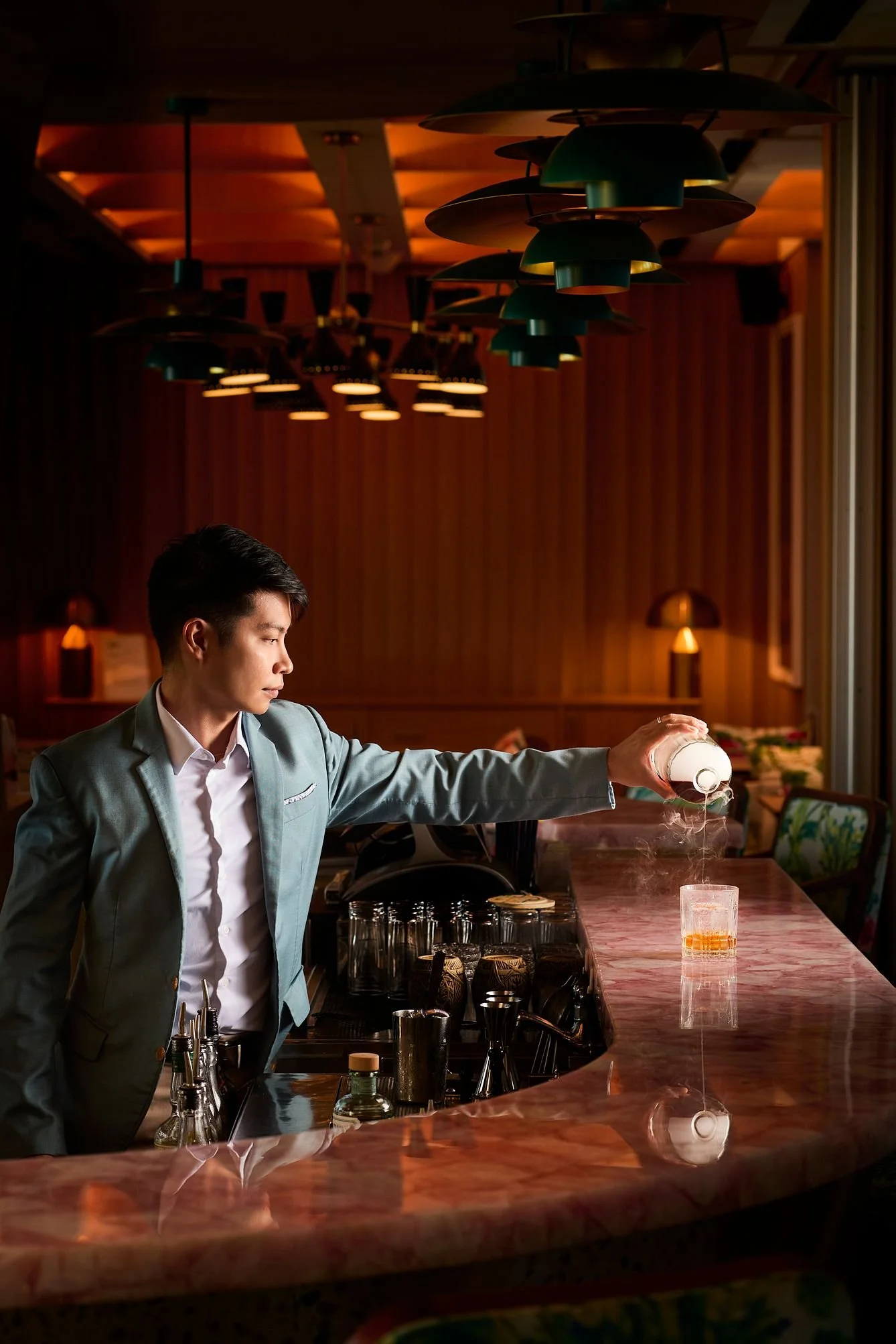 A bartender in a gray suit is pouring a drink at a bar counter in a dimly lit, stylish bar with wooden walls and ceiling, and decorative pendant lights overhead.