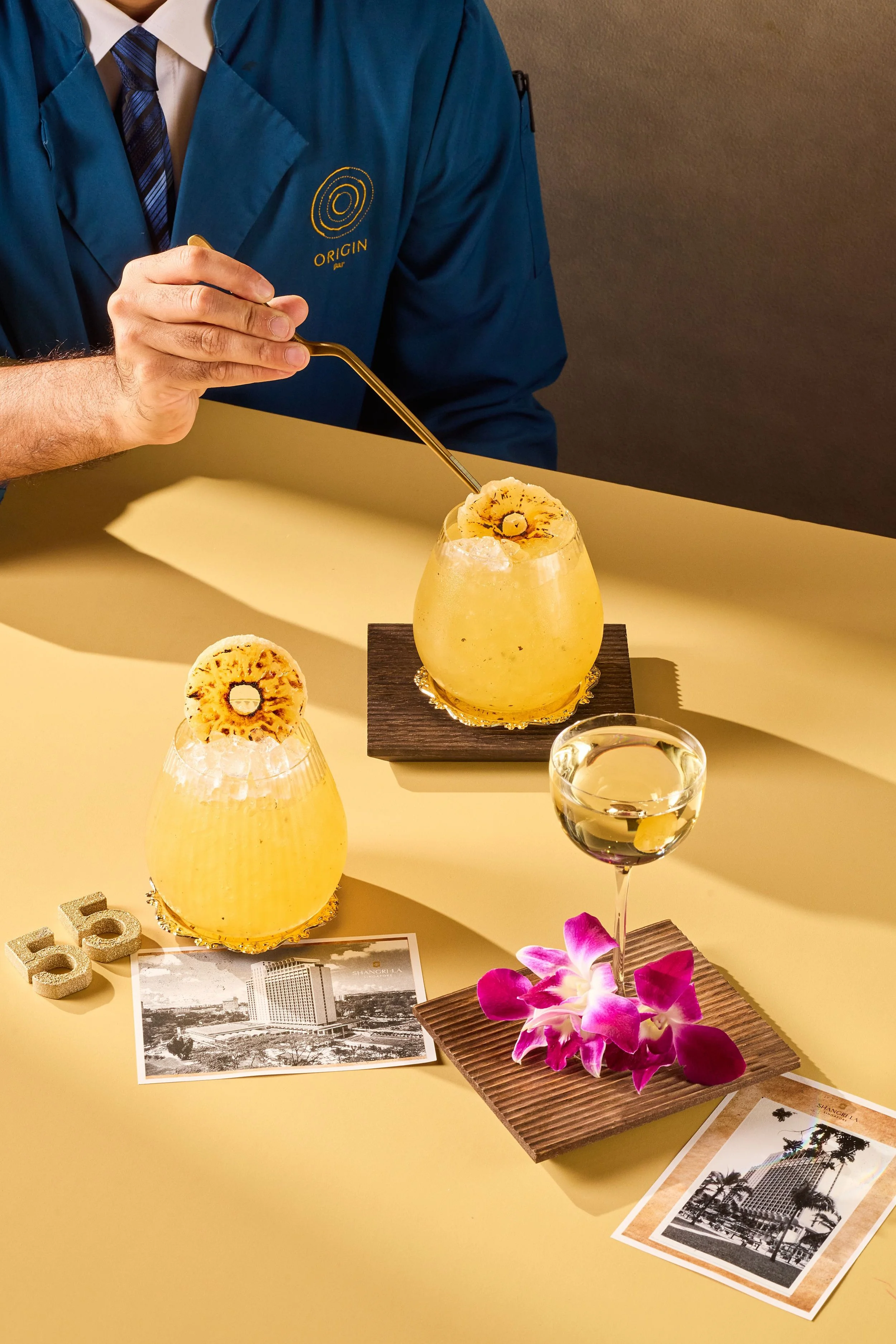 A person dressed in a blue blazer with a logo on the chest, sitting at a table with tropical drinks garnished with dried pineapple slices, a glass of red wine, and a small bouquet of purple orchids on a wooden mat. There are also postcards and decorative elements on the table, and the background has a plain wall.
