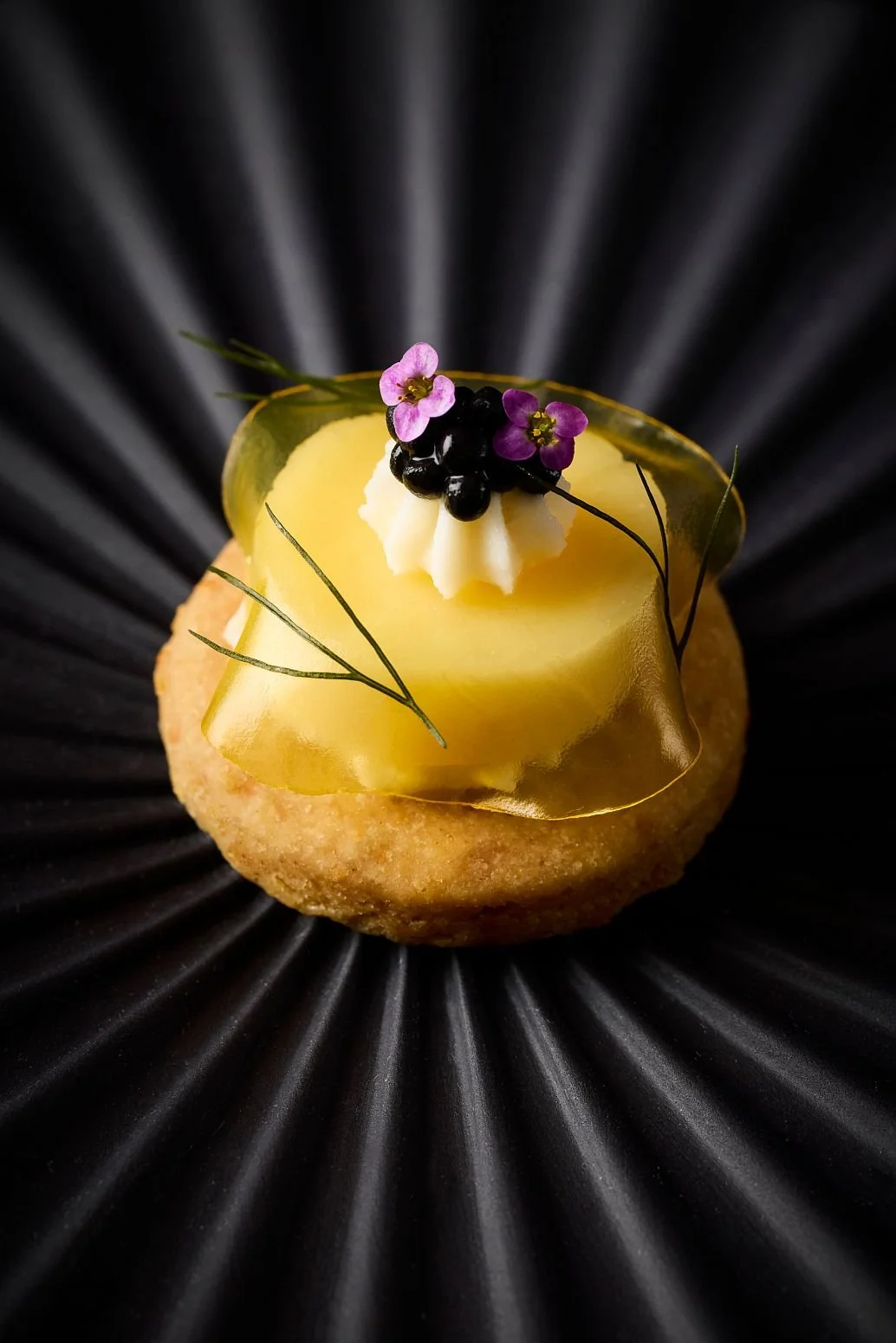 A close-up of a gourmet dessert with a round, golden cookie base topped with yellow gelatin, a dollop of white cream, blackberries, pink edible flowers, green leaves, and thin black edible sticks, on a black textured plate.